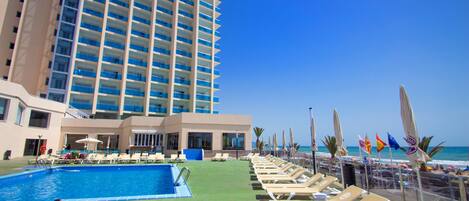Zona de piscina de hotel con sillas de descanso y sombrillas, un cielo azul despejado y un edificio grande al fondo.
