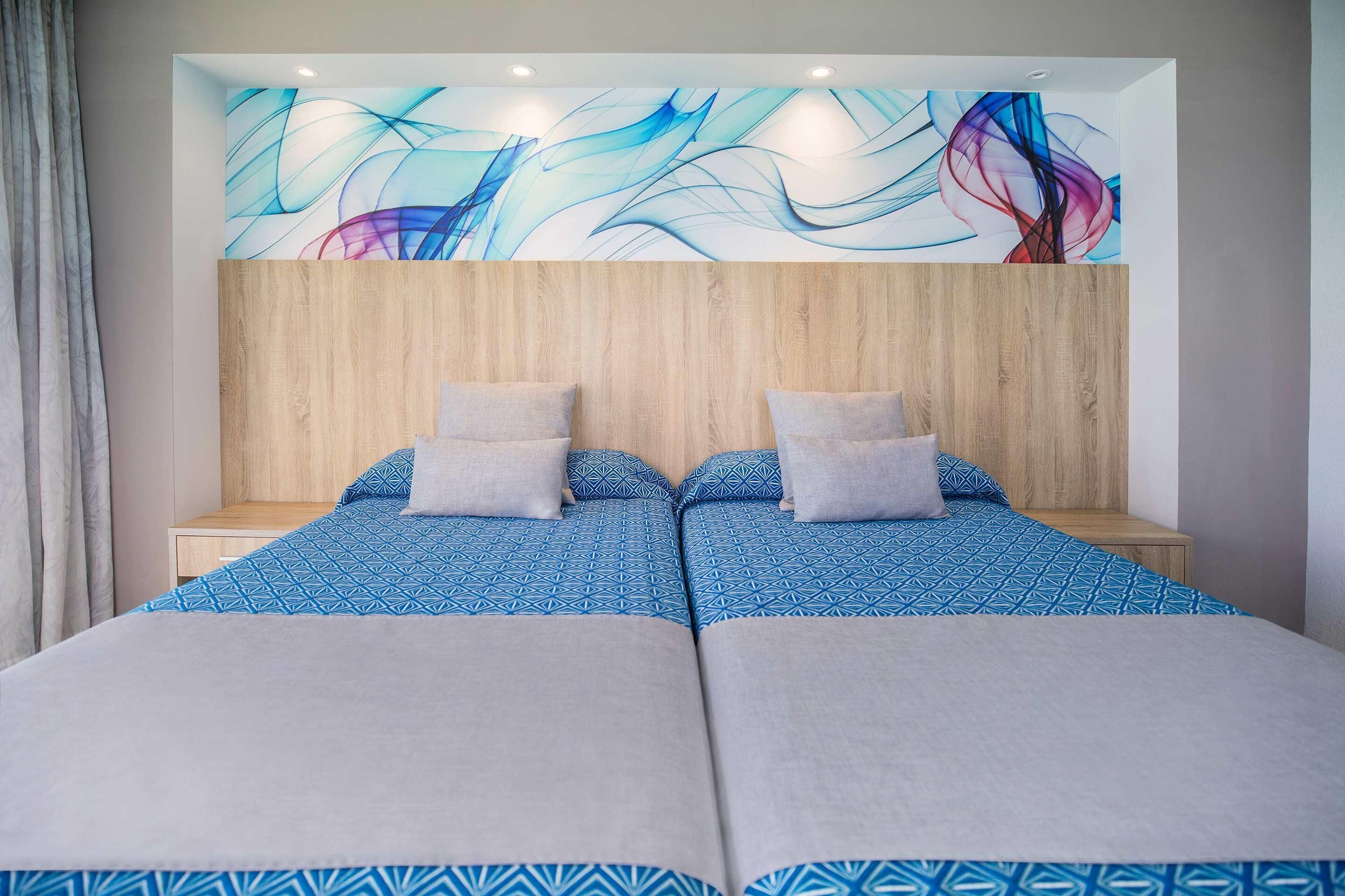 A double bed with a blue patterned bedspread and two pillows, a wooden headboard, and a wall with abstract art.