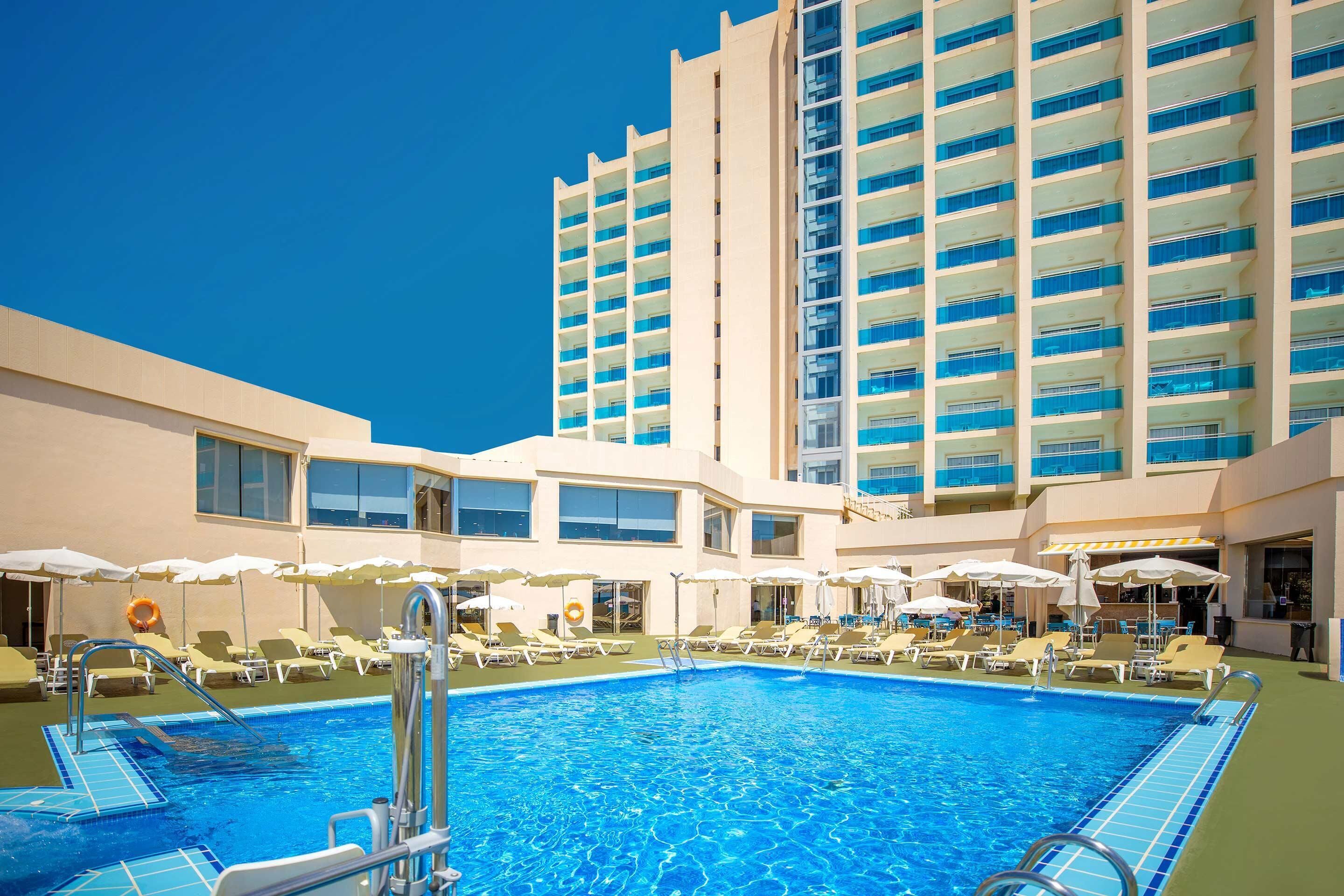 Una piscina con sillas de descanso y sombrillas, rodeada por un edificio de hotel con varios pisos y balcones.