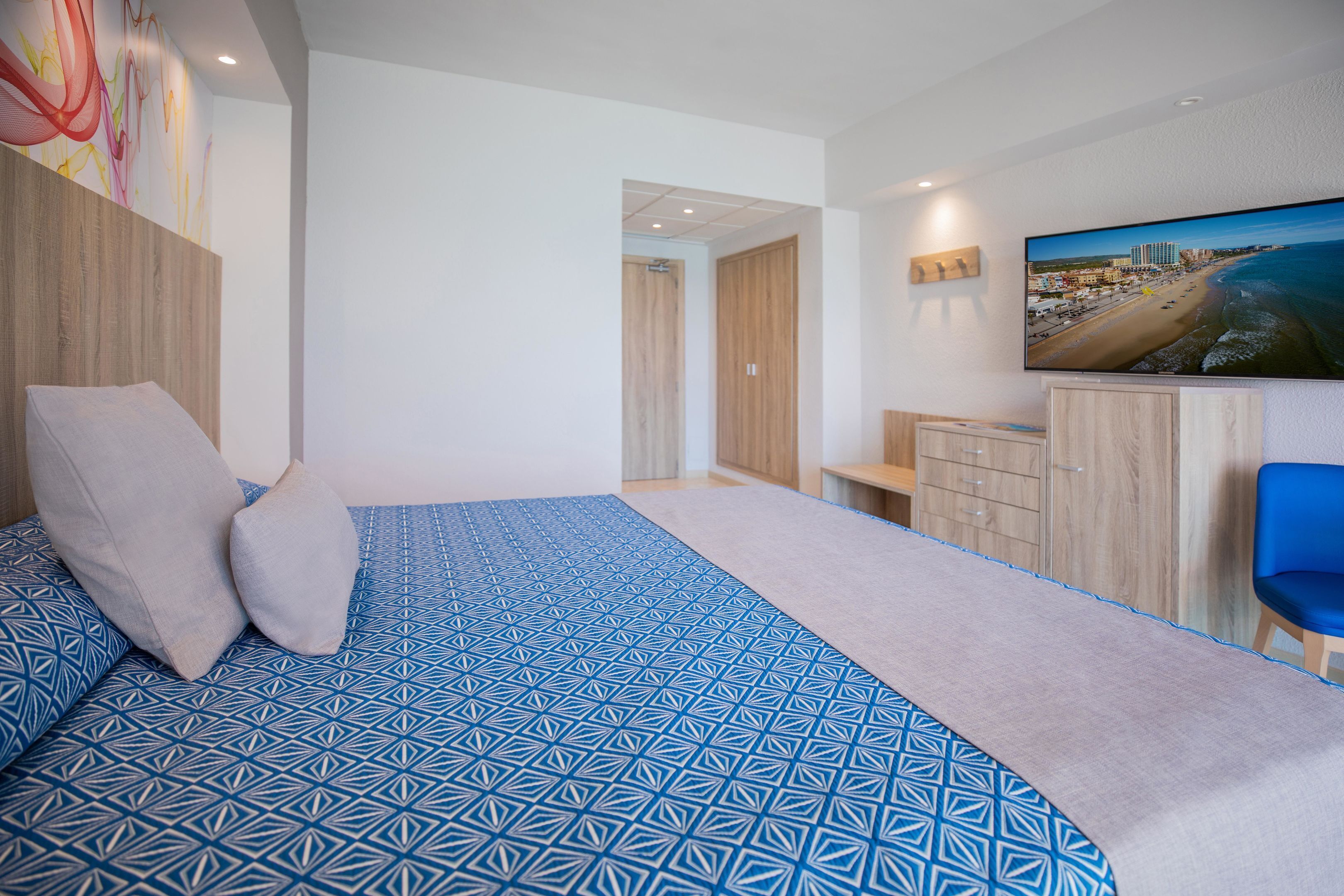 Una habitación de hotel moderna con una cama grande, un televisor montado en la pared y vistas a la playa.