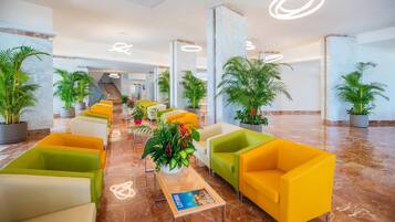 Een hotellobby met een marmeren vloer, rijen gele en groene stoelen, potplanten en een trap op de achtergrond.