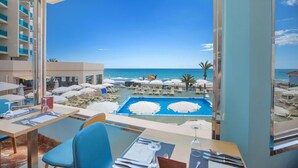 A hotel room with a view of a pool and beach umbrellas, a dining area with tables and chairs, and a balcony with a view of the ocean. - Hotel Servigroup Koral Beach (Oropesa del Mar)