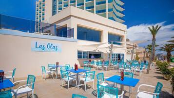 An outdoor patio area with tables and chairs, a sign reading "Las Olas," and a modern building in the background.