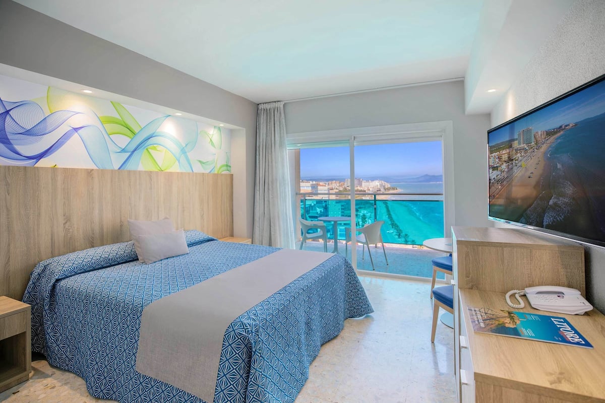 Habitación de hotel con cama, escritorio con teléfono y revista, televisión y balcón con vistas.