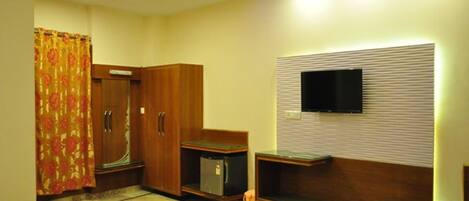 A hotel room with a wooden wardrobe, a small fridge, a TV mounted on the wall, and a bench.