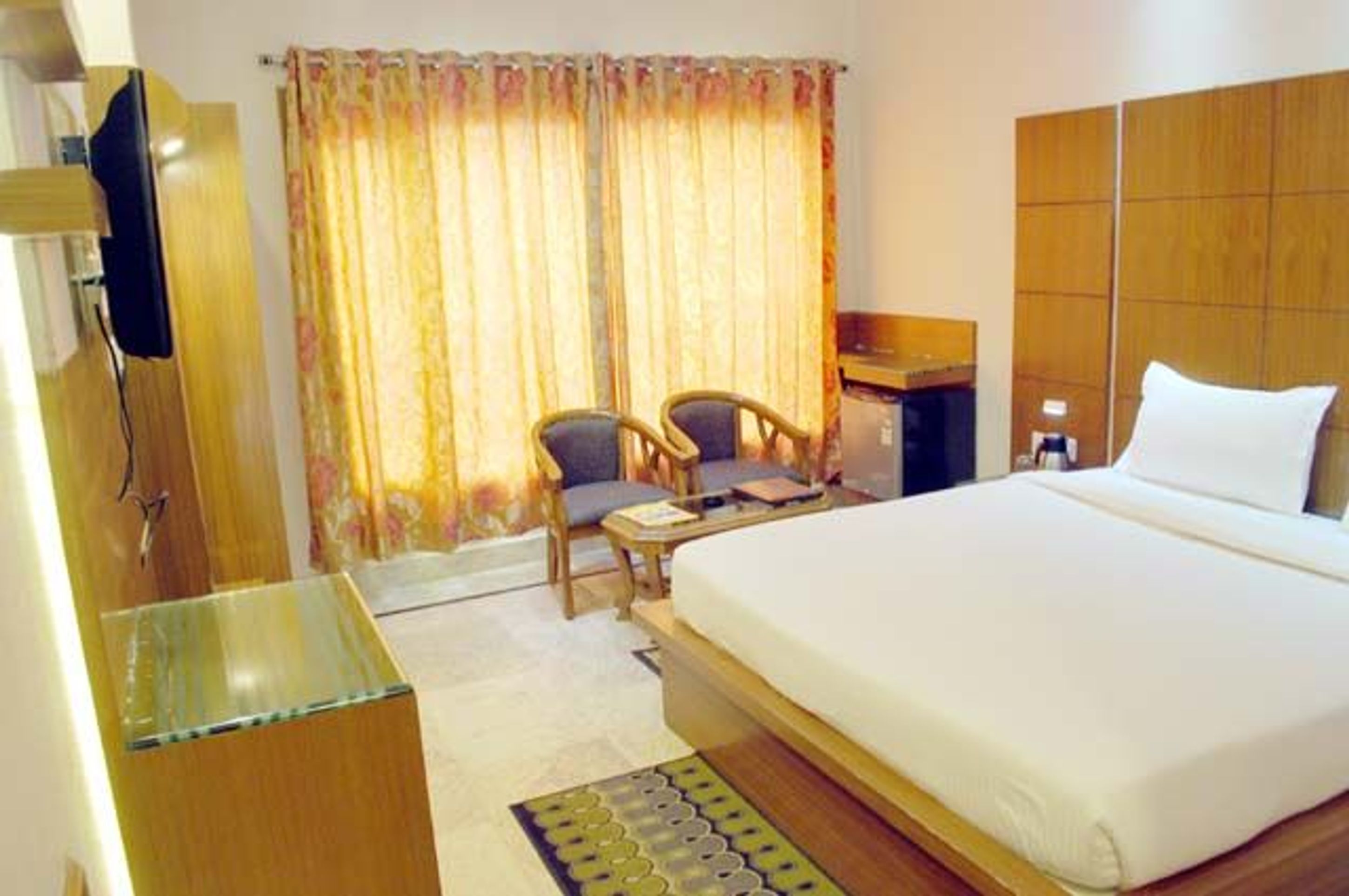 A hotel room with a large bed, a desk, two chairs, a television, and a window with curtains.