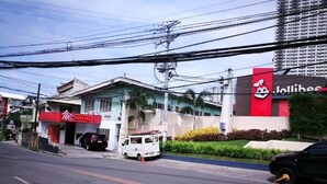 Un hostal con un letrero rojo, un restaurante Jollibee al otro lado de la calle y una camioneta blanca estacionada en la vía.