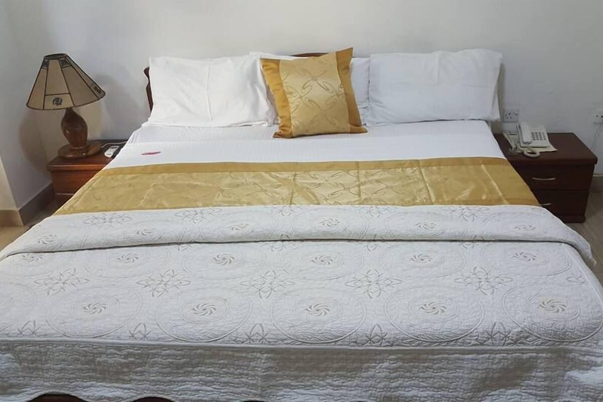A neatly made bed with a white and gold bedspread, a wooden nightstand with a lamp, and a phone on the side table.
