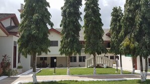 A two-story building with a symmetrical facade, a central entrance with a staircase, and a well-maintained lawn with evenly spaced trees. - Trust Lodge (Sekondi-Takoradi)