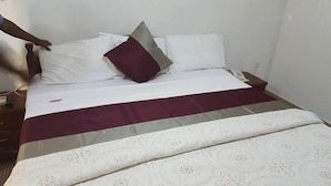 A neatly made bed with a white and maroon bedspread, a single maroon pillow, and a white pillow. - Trust Lodge (Sekondi-Takoradi)