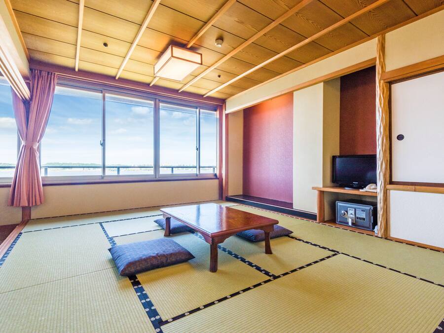 A traditional Japanese room with tatami flooring, a low wooden table, cushions, a television on a shelf, and a view through large windows.