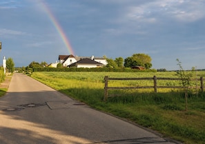 A rural road with a rainbow, a white house, and a wooden fence.