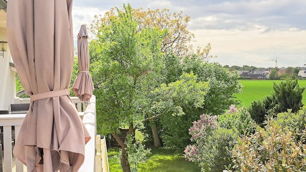 A balcony with a beige curtain and a white chair, overlooking a green garden with trees and shrubs.