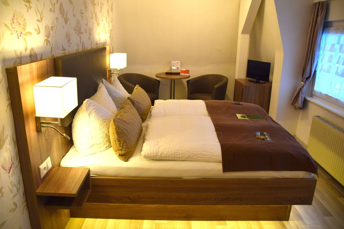 Una habitación de hotel con una cama, mesitas de noche, un televisor y una pequeña mesa con una lámpara.
