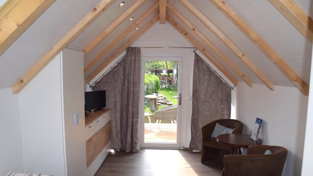 A cozy room with a sloped ceiling, a small kitchenette, a TV, a dining area with two chairs, and a sliding glass door leading to an outdoor patio. - Pension Lachmann (Koenigstein)