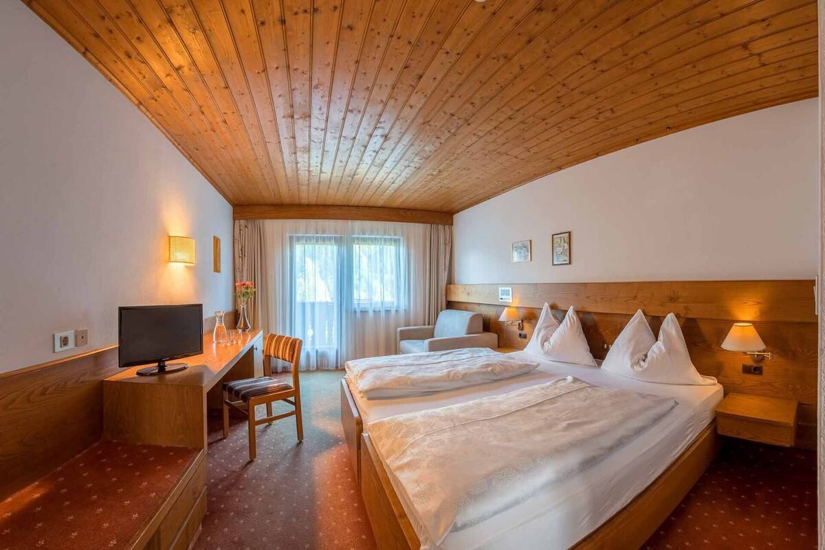 A hotel room with a wooden ceiling, a bed with white linens, a desk with a computer, a chair, and a window with curtains.