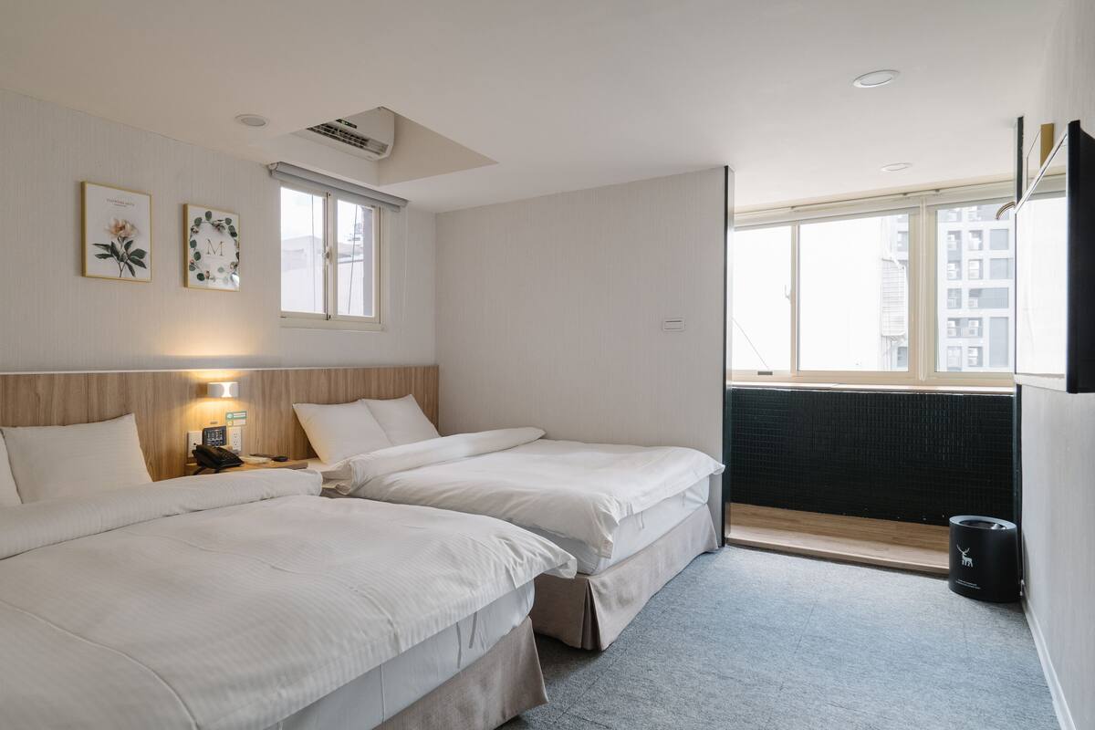 A hotel room with two beds, a bedside table with a lamp, a window with a view of buildings, and a wall-mounted air conditioner.