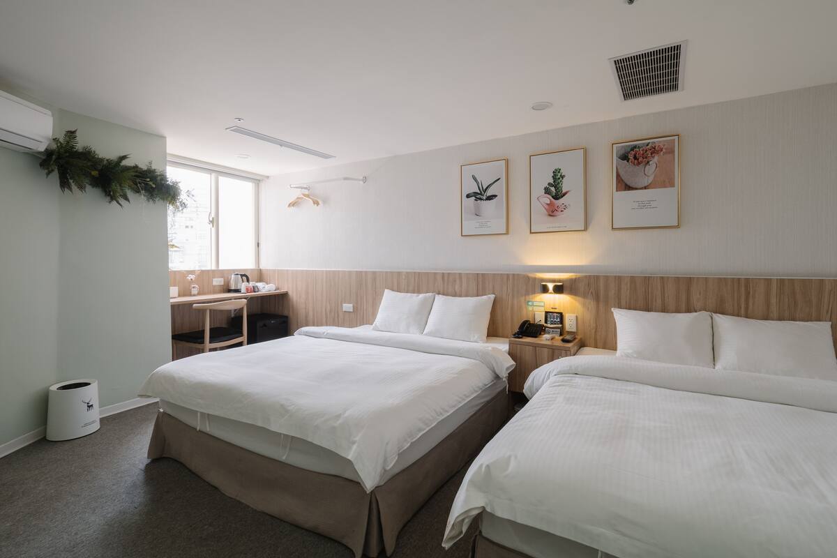 A hotel room with two beds, a desk, and a chair. There are framed pictures on the wall and a plant on the window sill.