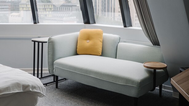 Una habitación de hotel moderna con un sofá azul claro, una pequeña mesita de madera y vistas a un paisaje urbano a través de grandes ventanales.