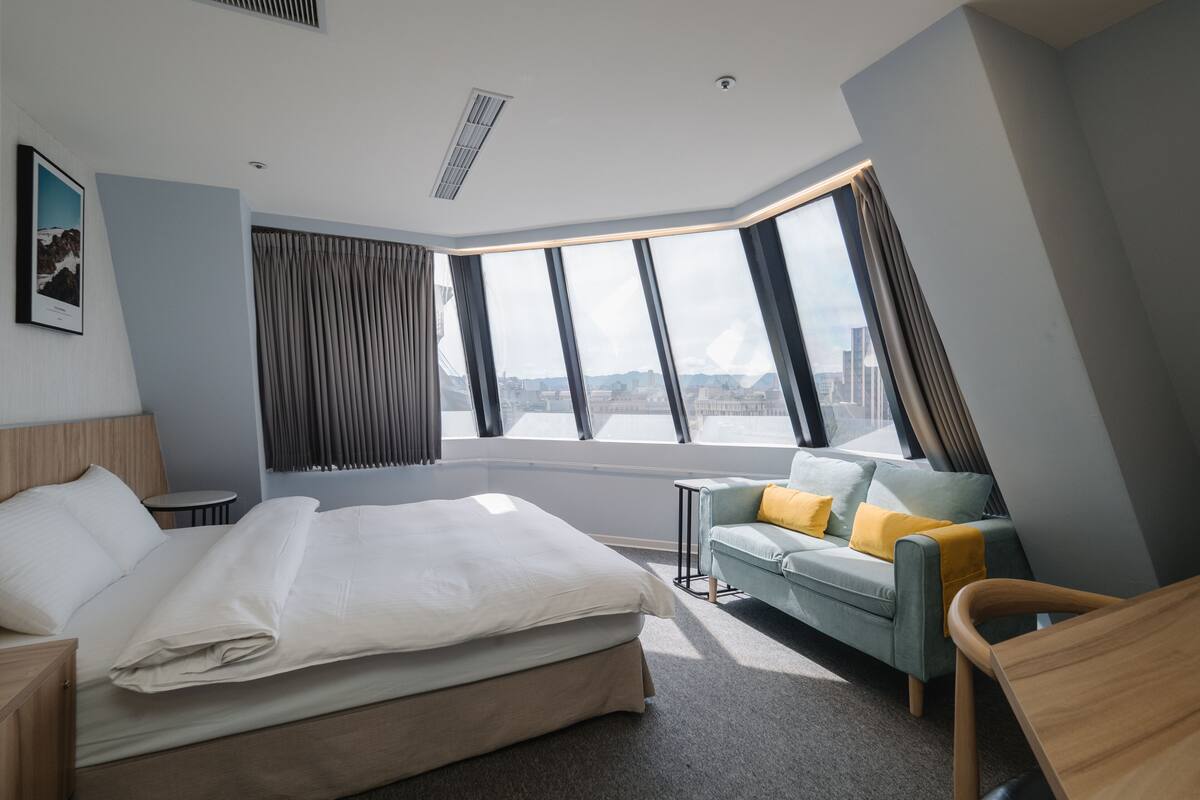 A modern hotel room with a large bed, a sofa, a small table, and a view of the city through large windows.