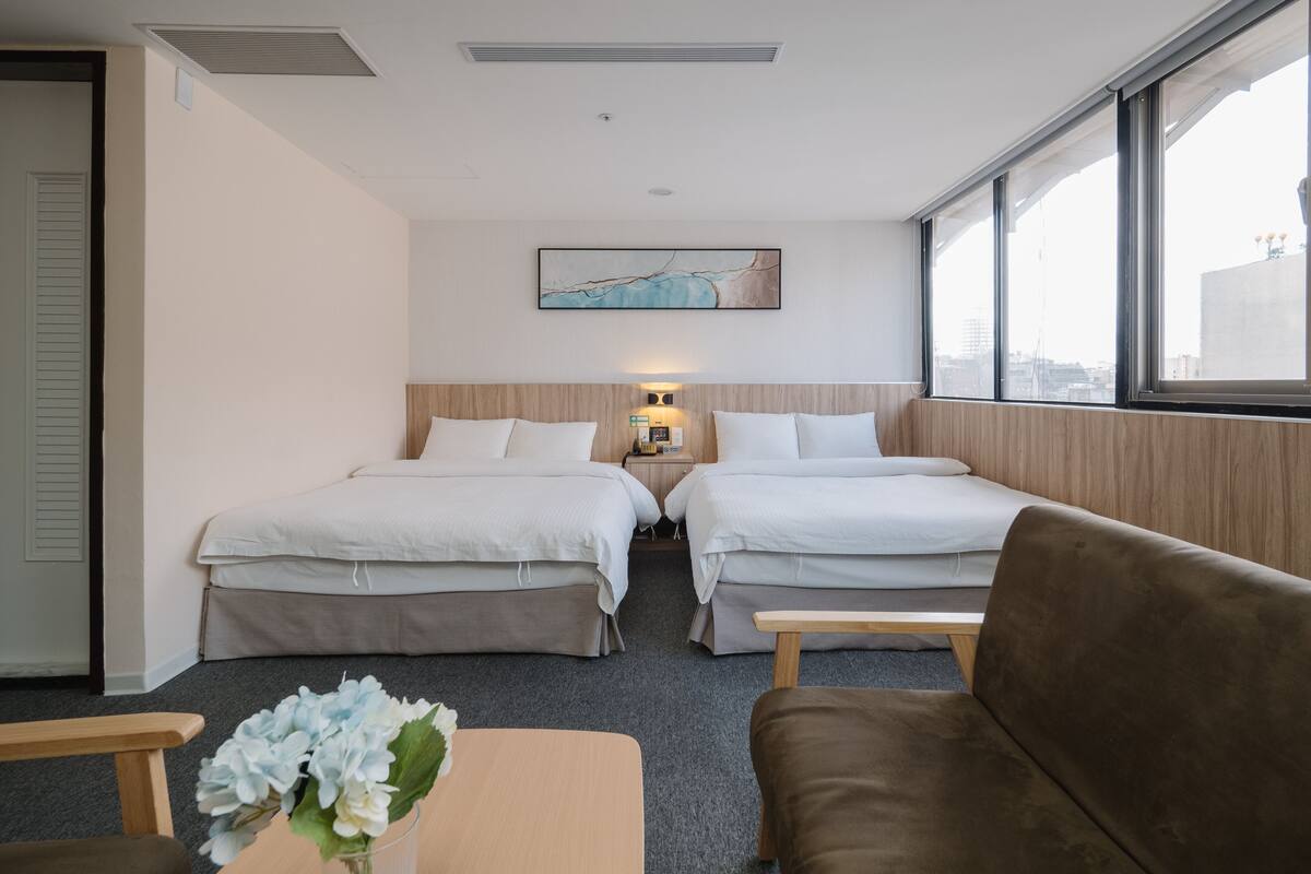 A hotel room with two beds, a sofa, a small table, a vase of flowers, and a city view through large windows.