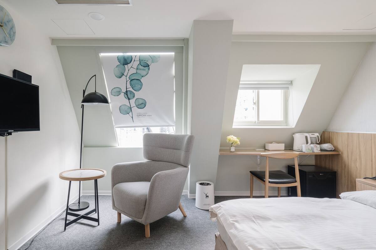 A modern hotel room with a bed, a desk with a chair, a TV, a window with a view, and a radiator.