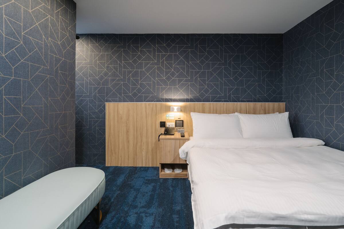 A hotel room with a bed, bedside table, and a small wooden nightstand with a lamp.
