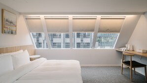 Una habitación de hotel moderna con una cama grande, una mesita redonda pequeña, un escritorio con una silla y un ventanal amplio con persianas.