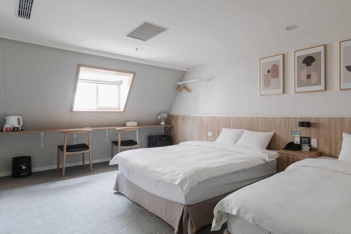 A hotel room with two beds, a desk with chairs, a window, and framed artwork on the wall.