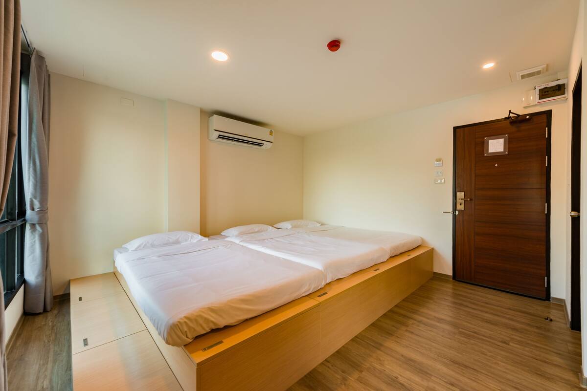 A single bed with a wooden platform, a window with curtains, an air conditioning unit, a wooden door, and a ceiling light.