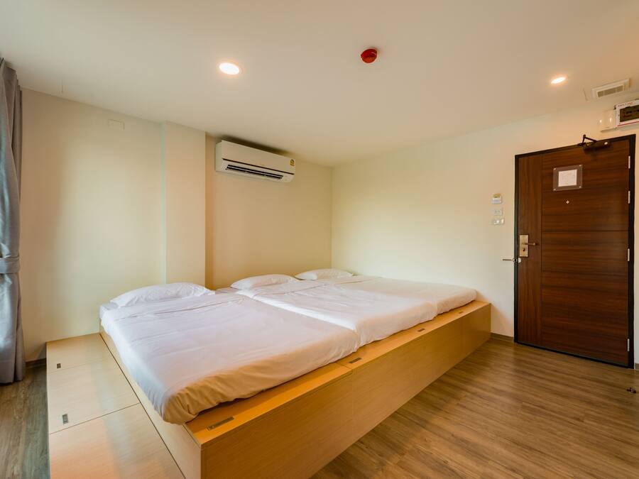 A single bed with a wooden platform, a window with curtains, an air conditioning unit, a wooden door, and a ceiling light.