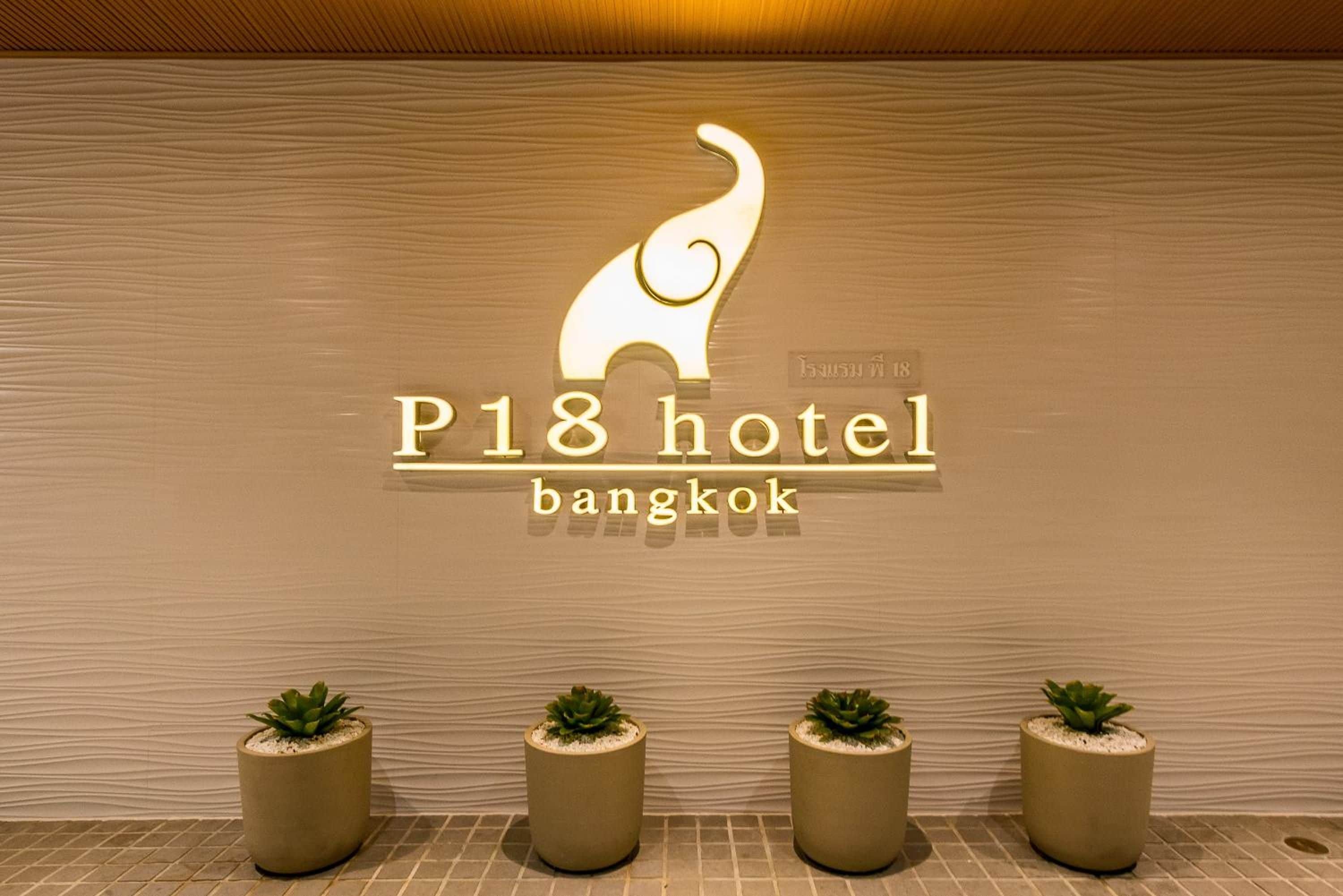A hotel sign with a stylized elephant logo and the name 'P18 hotel bangkok' against a textured wall. There are three potted plants in front of the sign.