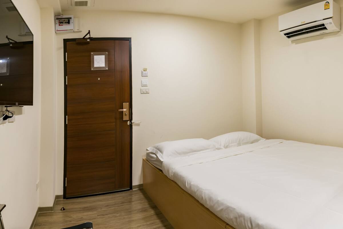 A single bed in a room with a wooden door, an air conditioning unit, and a small desk with a chair.