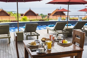 Eine Holzterrasse mit einem Esstisch, gedeckt mit Tellern, Tassen und Getränken, mit Blick auf einen Pool mit Liegestühlen und Sonnenschirmen.