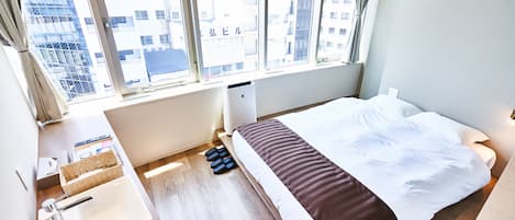 A hotel room with a bed, a bedside table with a lamp, a window with a view of buildings, and a small bathroom area.