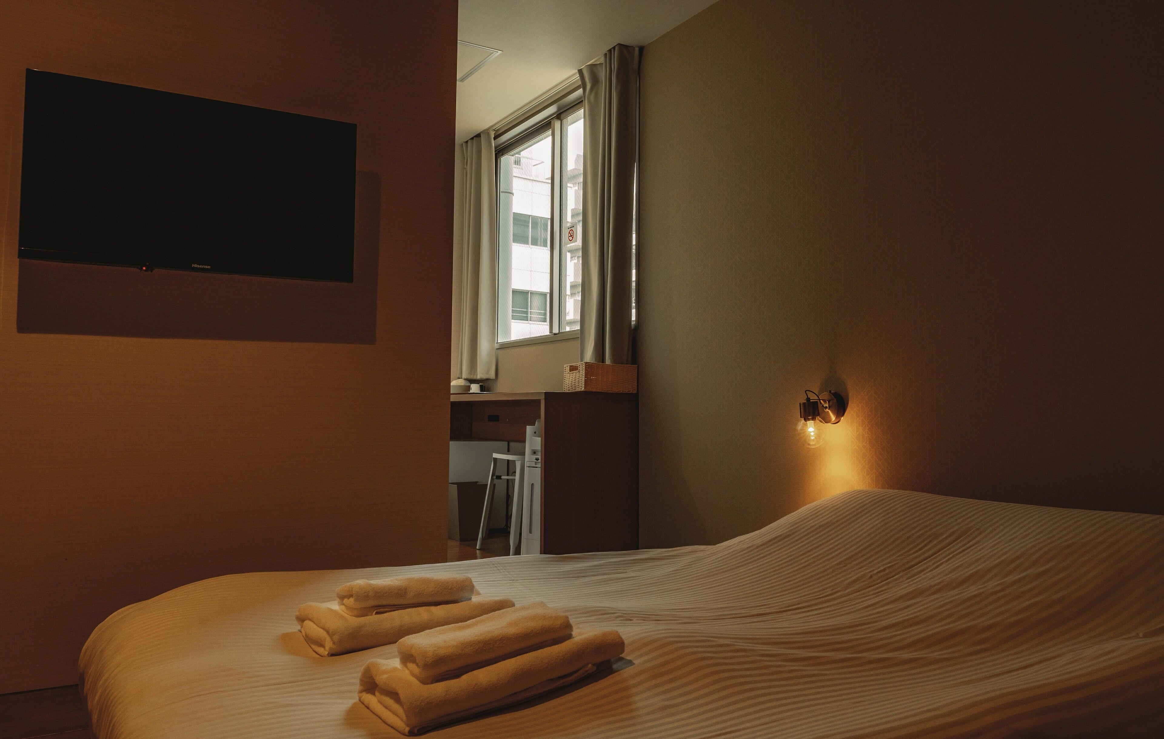 Ein Hotelzimmer mit Bett, Flachbildfernseher an der Wand, Fenster mit Vorhängen und einer Wandleuchte.