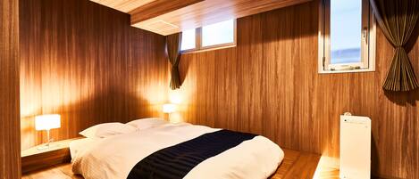 Una habitación con paneles de madera, una cama, dos lámparas de noche, un ventanal con cortinas y un termostato blanco en el suelo.