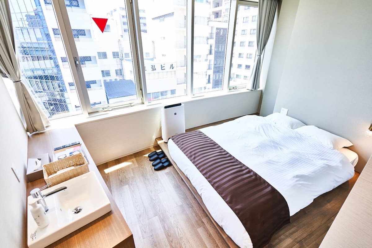 Et hotelværelse med en seng, et lille natbord med en lampe, et vindue med udsigt til bygninger og en radiator.