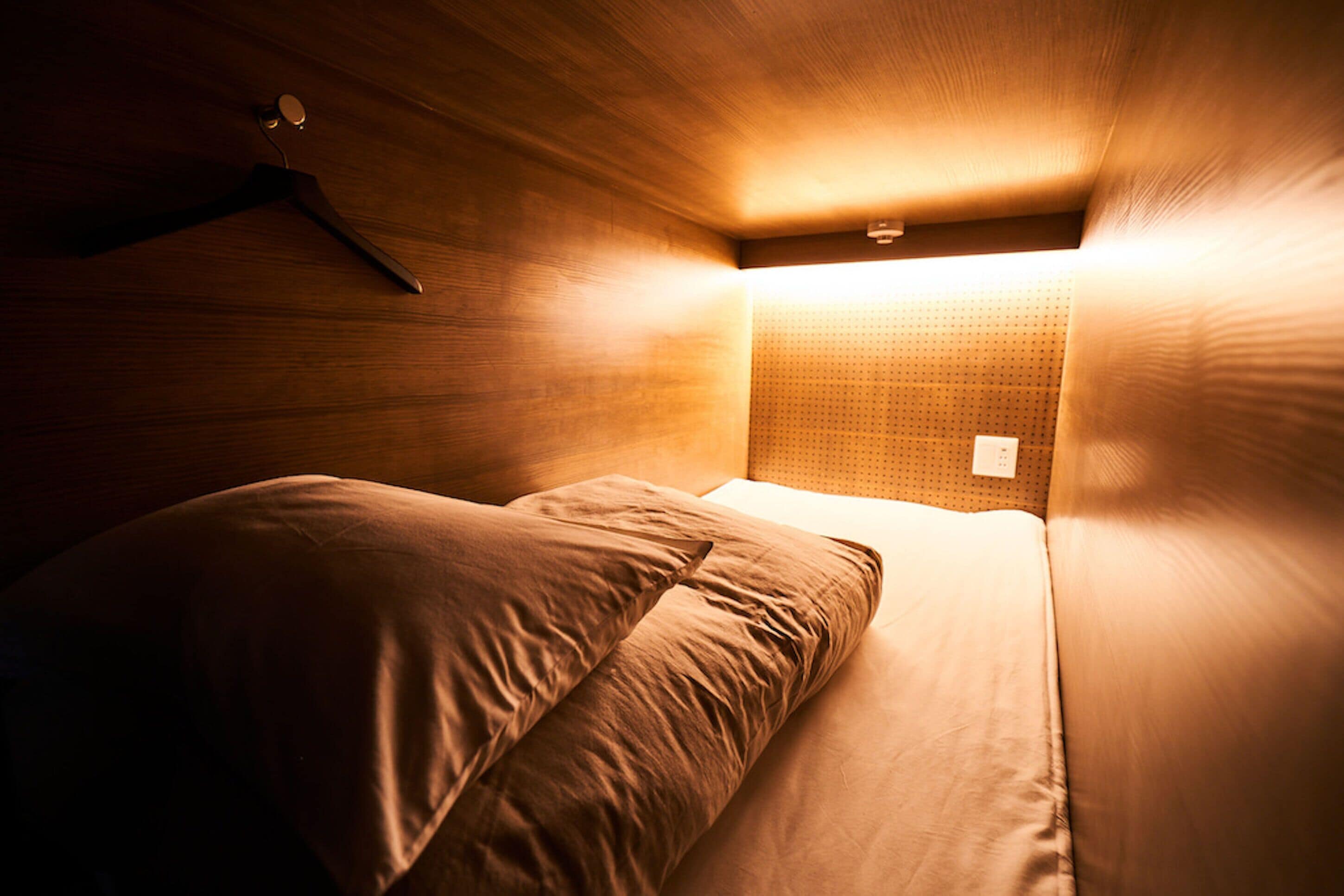 Ein kleiner, schummrig beleuchteter Raum mit einem Einzelbett, einem an der Wand hängenden Kleiderhaken und einer Wandleuchte.