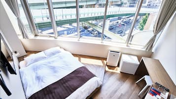 A hotel room with a bed, a desk, a chair, a small table, and a view of an urban area with buildings and a bridge.