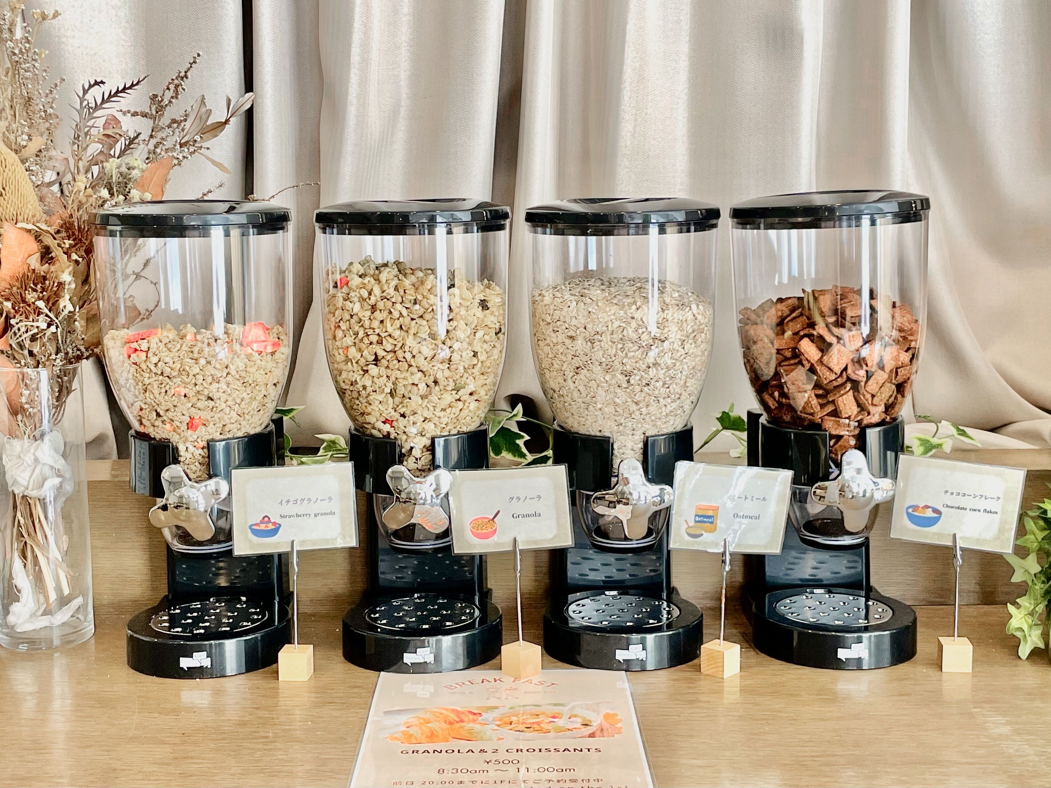Quatre distributeurs de granola, chacun muni d’une étiquette indiquant le type de granola qu’il contient, un menu proposant différentes sortes de granola et de croissants, et un vase décoratif contenant des fleurs séchées.