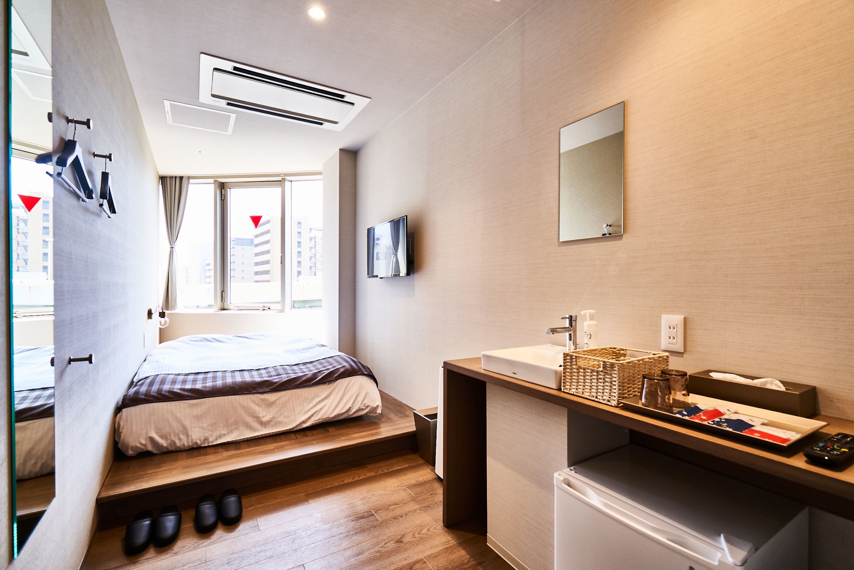 A modern hotel room with a bed, built-in storage, a small kitchenette, and a large window offering a city view.