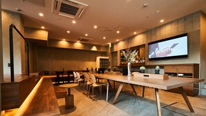 Un hall d’hôtel moderne avec un long comptoir d’accueil, un espace salon et un téléviseur diffusant le mot « AND HOSTEL ».