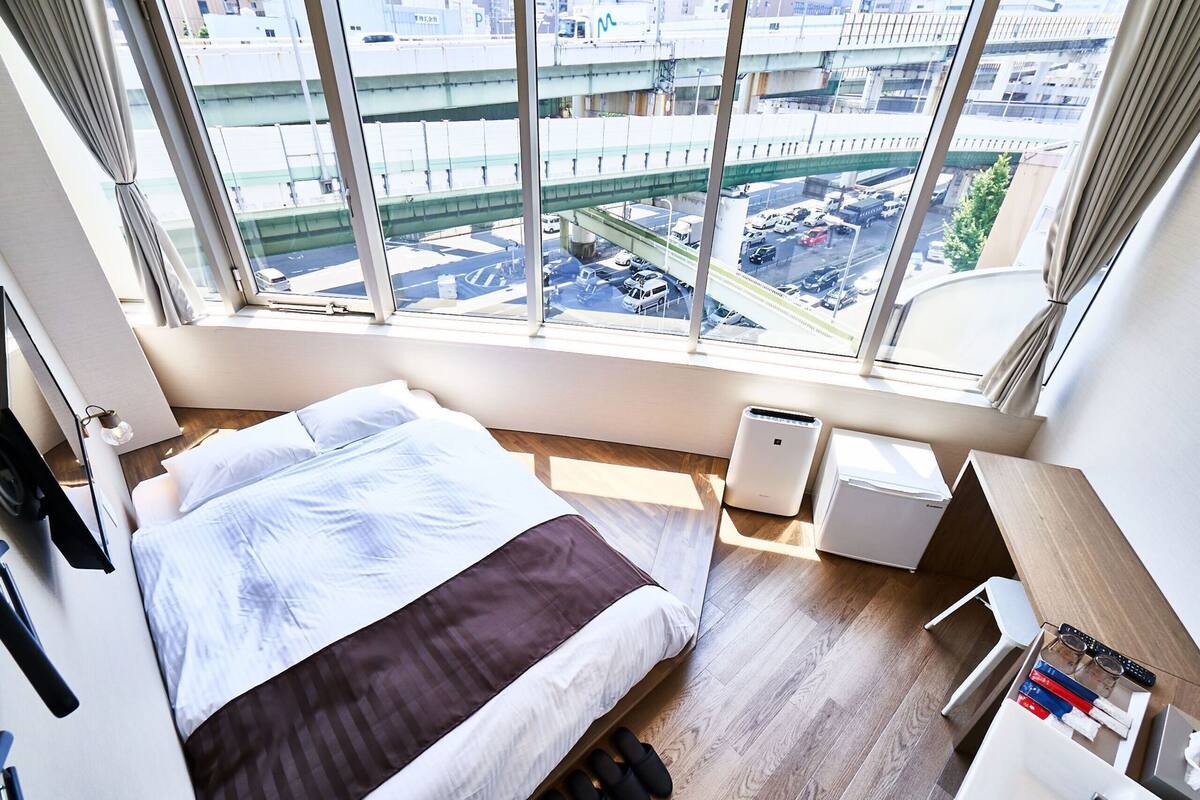 Et hotelværelse med en seng, et lille skrivebord, en varmeblæser og udsigt til en travl gade og motorvej.