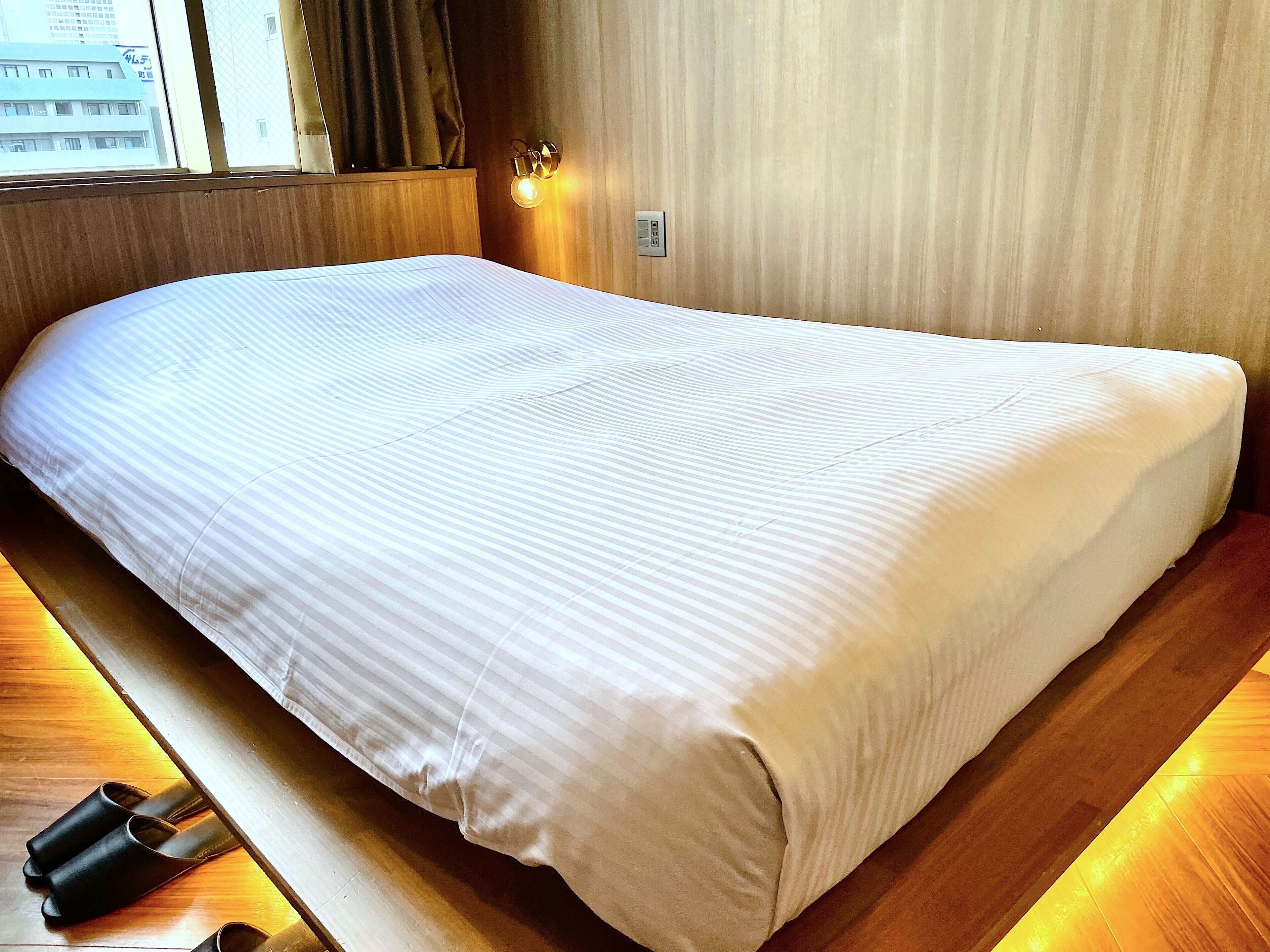 A neatly made bed in a room with wooden flooring and a window with curtains.