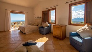 A spacious room with a bed, two sofas, a wooden cabinet, and large windows offering a mountain view. - Villa Noelia (Arzachena)