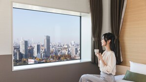 A hotel room with a large window offering a city view, a bed with white bedding, and a green pillow. - Hotel Crown Hills Ueno Premier (Tokyo)