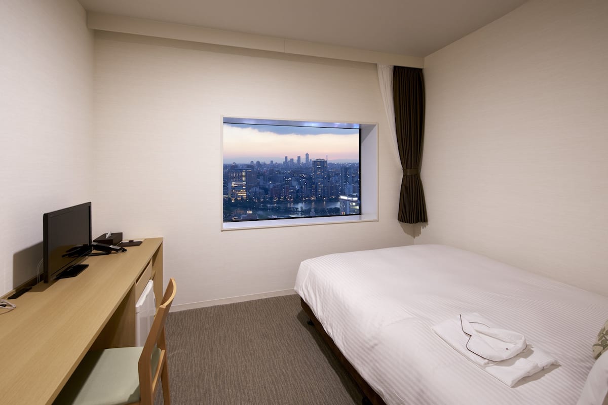 Een hotelkamer met een bed, een bureau met een computer, een stoel, een raam met uitzicht op de stad en een gordijn.