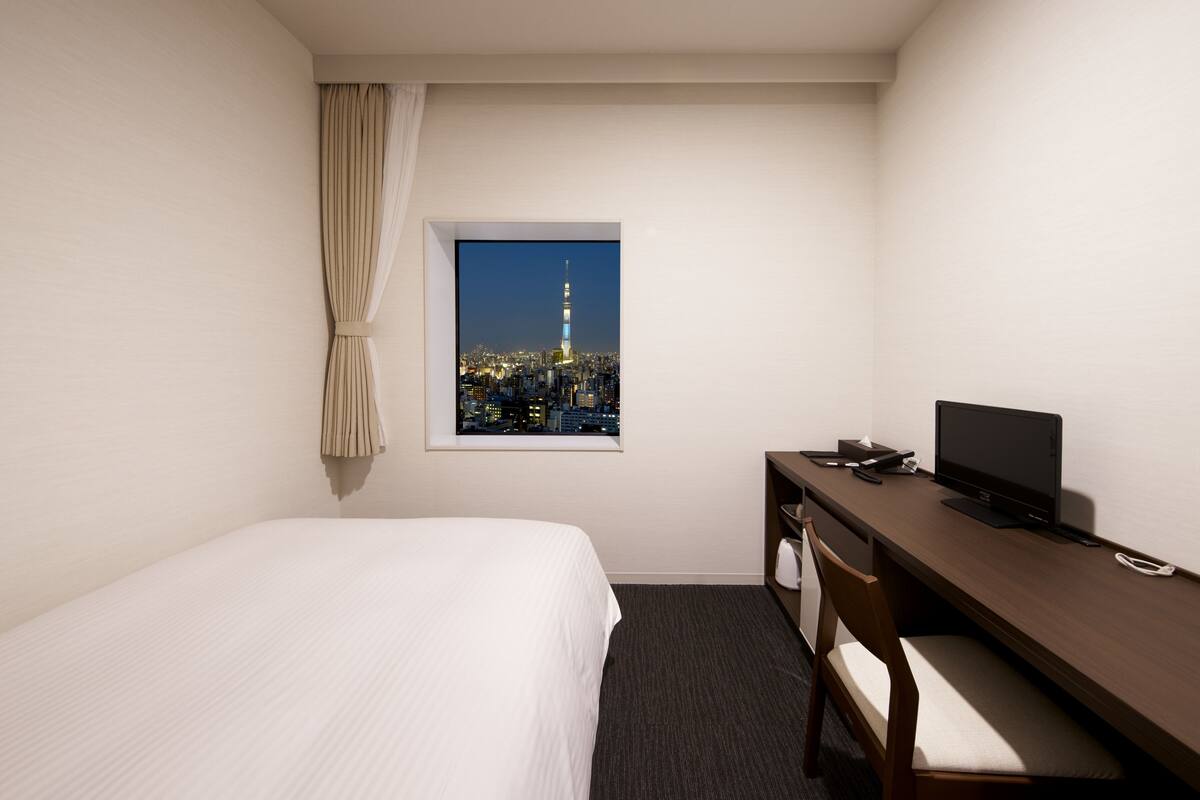 Een hotelkamer met een bed, een bureau met een computer, een stoel, een raam met uitzicht op de stad en een gordijn.