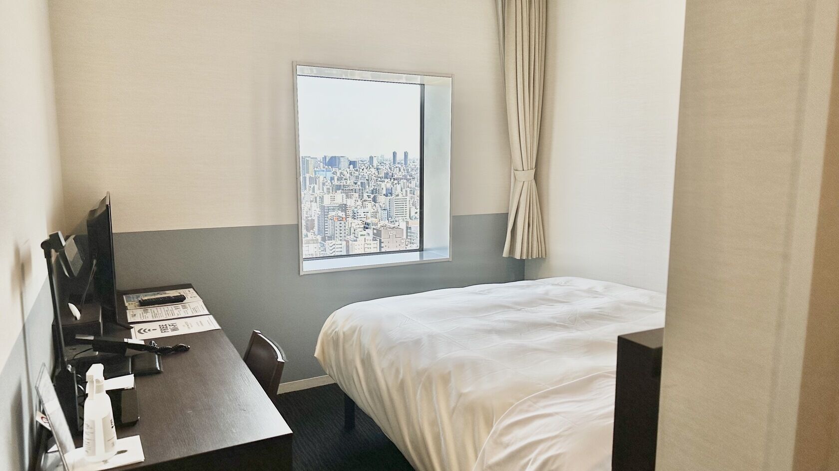 A hotel room with a bed, a desk with a computer, a window with a cityscape view, and a curtain.